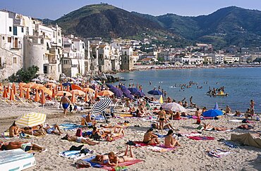 ITALY Sicily Palermo Cefalu. View across busy sandy beach with sunbathers on sand and swimming in the sea overlooked by waterfront apartments and hills