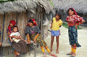 PANAMA San Blas Islands Kuna Indians Kuna Indian family with women wearing brightly coloured tradtional mola panel blouses or dulemolas outside their thatched home  their legs bound with traditional design strings of coloured glass beads Cuna Caribbean  house American Central America Colored Female Woman Girl Lady Hispanic Kids Latin America Latino Panamanian West Indies