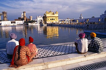 Sikh men woman and child sitting on marble walkway beside the sacred pool looking towards the Golden Temple reflected in the water, Amritsar, Punjab, India