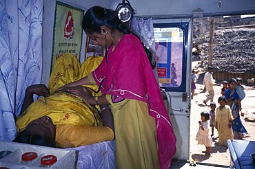 Female health worker examining a pregnant woman in a mobile clinic, Children and woman seen outside through open door, Bangalore, Karnataka, India