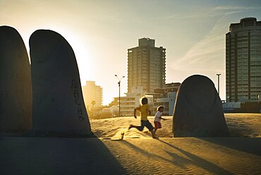 La Mano Sculpture of a hand on the beach at Praya Brava with children playing in the evening light, Punte del Este, Uruguay