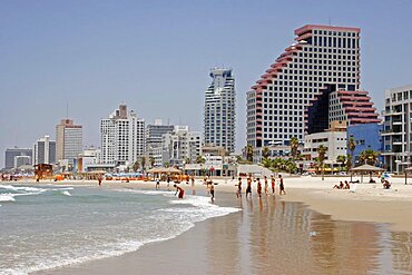 Beach with people sunbathing and at waters edge overlooked by high rise modern city buildings reflected on wet sand, Tel Aviv, Israel