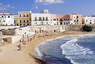 View of old city with traditional houses beside sea wall overlooking curving sandy beach with people sunbathing and swimming, Gallipoli, Puglia, Italy