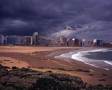 High rise city buildings overlooking beach with people in water on beach and surfing, Grey cloudy sky with approaching storm, Rocks and seaweed in foreground, Gijon, Asturias, Spain