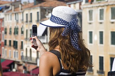 Italy, Veneto, Venice, Young female tourist in white straw hat taking photograph on mobile phone from Rialto bridge looking along Grand Canal