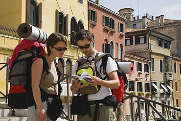 Italy, Veneto, Venice, Two young backpacker tourists wearing sunglasses and carrying rucksacks, rolled up sleeping mats and moneybelts, consulting guide book while standing on canal bridge in Centro Storico centre of Venice