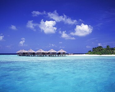 MALDIVES  Baros View across expanse of turquoise sea towards thatched beach huts on stilts  beach and tropical vegetation.