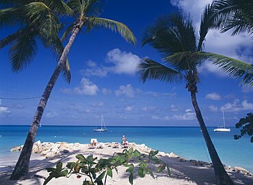 WEST INDIES Antigua Dickenson Bay Sandy beach and rocks with people sunbathing on loungers and man fishing.  Aquamarine water and yachts framed by palms in the foreground.