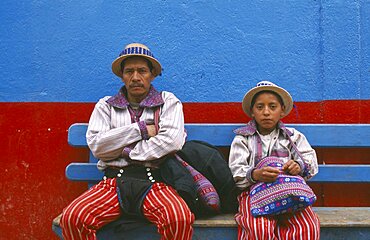 GUATEMALA  Todos Santos Cuchumatan Father and Son dressed identically  sitting on a blue bench against a blue and red wall.