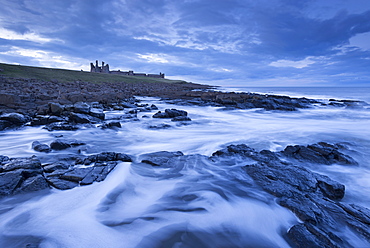 Waves swirl around the rocky shores below Dunstanburgh Castle at twilight, Northumberland, England, United Kingdom, Europe