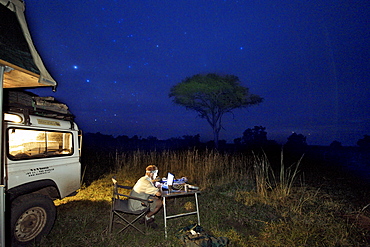 A night time view of a man working at a laptop alongside a Land Rover and tent pitched in a game park.