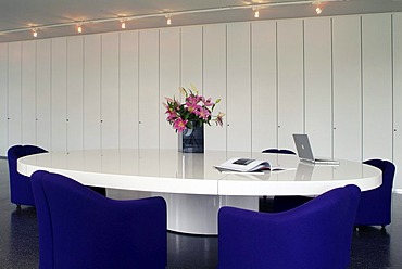 Oval conference table with a laptop and chairs in a modern design in front of a white office wall unit