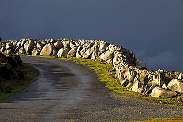 Sunlit stone wall fence and road with dark storm cloud, Republic of Ireland, Europe