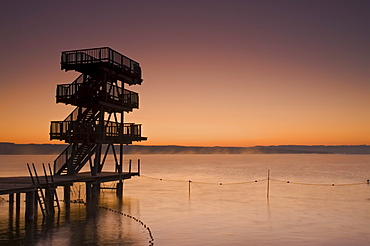 The diving platform at the shore of the calm Ammersee lake in front of a colorful sunrise, Utting, Bavaria, Germany, Europe