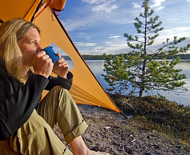 Woman sitting in front of a tent, looking into the distance and drinking out of a cup, Femundsmarka National Park, Femundsmark, Norway