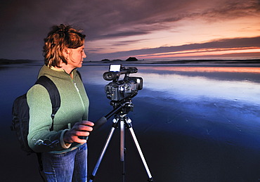 Camerawoman with movie camera filming sunset with a tripod at Meyers Creek Beach, Pistol River State Park, Oregon Coast, Oregon, USA, North America