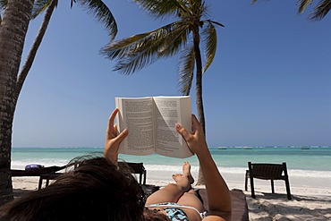 Girl, 16 years, reading a book on the beach under palm trees