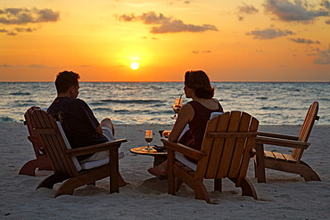 Couple sitting on chairs at the beach with long drinks in front of a golden sundown, Rihiveli, Island, Maldives, South Male Atoll, Archipelago, Indian Ocean, Asia