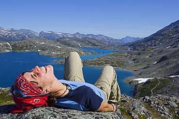 Young woman laughing, lying on a rock, relaxing, hiker enjoying the panorama on summit of historic Chilkoot Pass, Chilkoot Trail, Crater Lake behind, alpine tundra, Yukon Territory, British Columbia, B. C., Canada