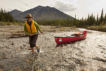 Young woman lining, wading, pushing, pulling a canoe in shallow water, Wind River, Peel Watershed, Northern Mackenzie Mountains behind, Yukon Territory, Canada