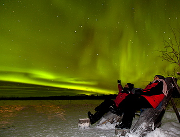 Young couple, woman and men, sitting in a wooden chair, watching northern polar lights, pointing, Aurora Borealis, green, near Whitehorse, Yukon Territory, Canada