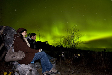 Couple, woman and man, sitting in a wooden chair, watching swirling Northern lights, Polar Aurorae, Aurora Borealis, green, near Whitehorse, Yukon Territory, Canada