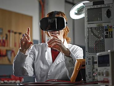 Woman with VR goggles and lab coat sitting in an electronics laboratory, Austria, Europe