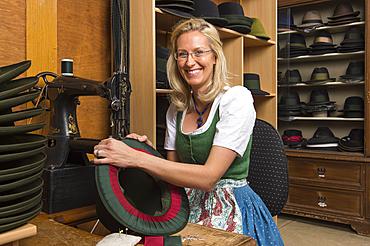 Woman sewing silk ribbon and inner lining on hat edge, finished hats on shelves behind, hatmaker workshop, Bad Aussee, Styria, Austria, Europe