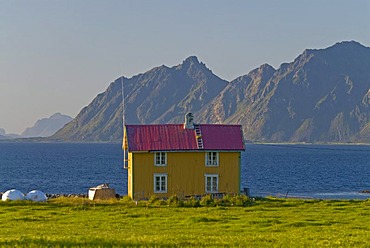 A yellow house in a green meadow near Sanden with steep mountains at back, Lofoten, Nordland, Norway, Europe