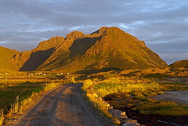 A gravel road winding towards a steep mountain illuminated by warm evening light in Sandoya, Nordland, Norway, Europe