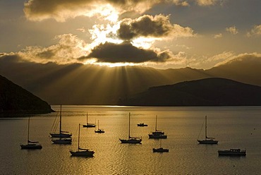 Sun beams over Akaroa Harbour and backlit boats in early morning light, Banks Peninsula, Canterbury region, South Island, New Zealand