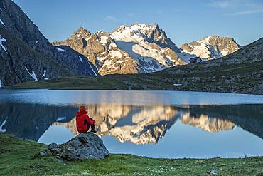 A hiker sitting on a rock contemplates the landscape at the edge of Lake Goleon (2438m) in the massif of Oisans with in the background the Meije (3983m) and the Rake (3809m), Ecrins National Park, Hautes-Alpes, France