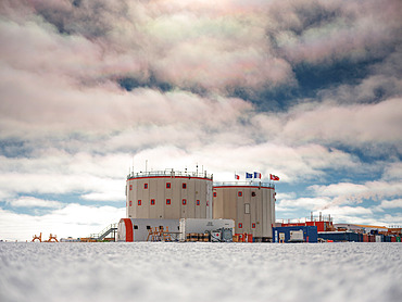 Concordia Station, Italian, European, French, Swiss and German flags. Tent, fire escape, containers, kerosene tanks, smoke from the power station, iridescent clouds. The station under a sky lightly covered with low, iridescent clouds. Concordia Antarctic Research Station, Dome C plateau, East Antarctica.