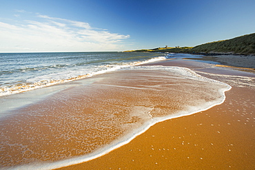 Waves on the beach at high tide near Low Newton by the Sea, looking towards Dunstanburgh castle, Northumberland, UK.