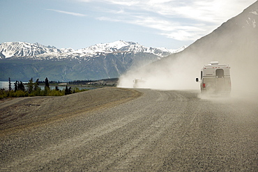 Following an RV along a lakeside gravel track, it's kicking up quite a dust cloud.  Set against snow capped mountains in the Alaskan Interior. The Interior, Alaska, USA