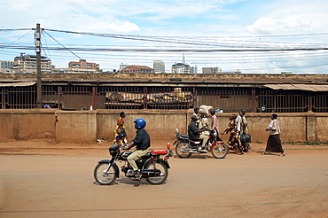 With the Kampala skyline appearing over the back wall of the Taxi park, pedestrians and motorcyclists share the roads.  Mopeds are a very popular and convenient form of transport around the City. . Kampala, Uganda, East Africa