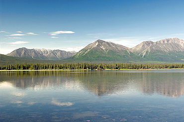 Clear blue sky with thin fluffy clouds over mountain range in the Alaskan Interior, leading down to the water's edge.  Lake bordered by dense forrest. The Interior, Alaska, USA