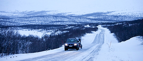 Car with trailer towing a snowmobile travels through arctic wilderness at nightfall by Kilpisjarvi on route from Norway into Finland