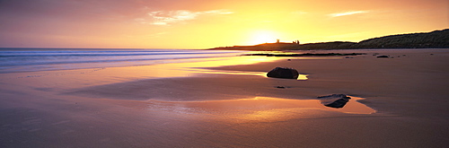 View along Embleton Bay at sunrise, with silhouette of Dunstanburgh Castle in the distance, near Alwick, Northumberland, England, United Kingdom, Europe