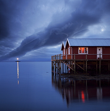 Rorbuer on stilts at dusk with lighthouse, Lofoten Islands, Norway, Scandinavia, Europe