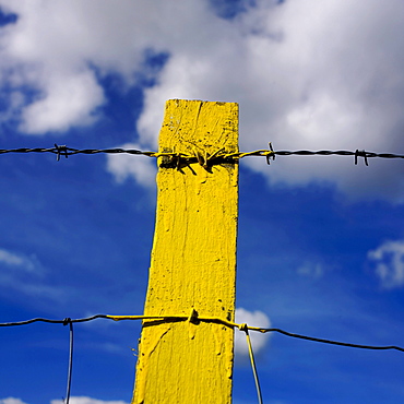 Yellow post, barbed wire, against blue sky with clouds