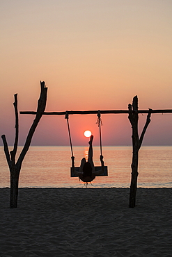 Silhouette of young woman on swing at Ong Lang Beach at sunset pretending to be kicking the sun like football, Ong Lang, Phu Quoc Island, Kien Giang, Vietnam, Asia