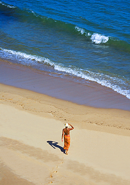 A woman dressed in a sarong with her back bare, walks in golden colored sand towards the water. Algarve, Portual.