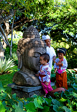 3 Balinese children playing in tradintional clothes around a huge Buddha statue, in a tropical setting.