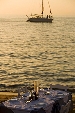 View over a table with glasses and plates to a sailingboat on the sea, Little Venice, Mykonos-Town, Mykonos, Greece