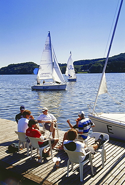 A group of people sitting on a jetty at a lake, sailing boats in the background, Baldeney lake, Essen, Ruhr Valley, Ruhr, Northrhine, Westphalia, Germany