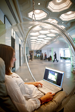 Woman sitting in a hanging ball chair while using a laptop, hotel lobby, Brussels, Belgium