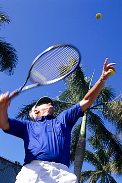 Upward view of senior man playing tennis, palm trees and blue sky 