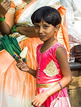 A young girl in a bright pink dress eating a popsicle and smiling for the camera, Mumbai, Maharashtra, India