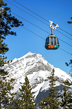 Gondola pod travelling along cables with snow covered mountain and blue sky in the background framed by evergreen trees, Banff National Park, Banff, Alberta, Canada